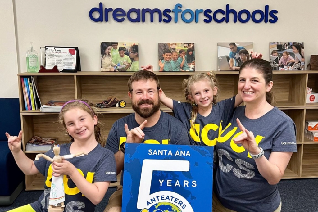 Four people wearing matching UCI shirts pose indoors in front of a “dreams for schools” sign, holding hand gestures beside a blue sign that reads “Santa Ana 5 Years Anteaters.”