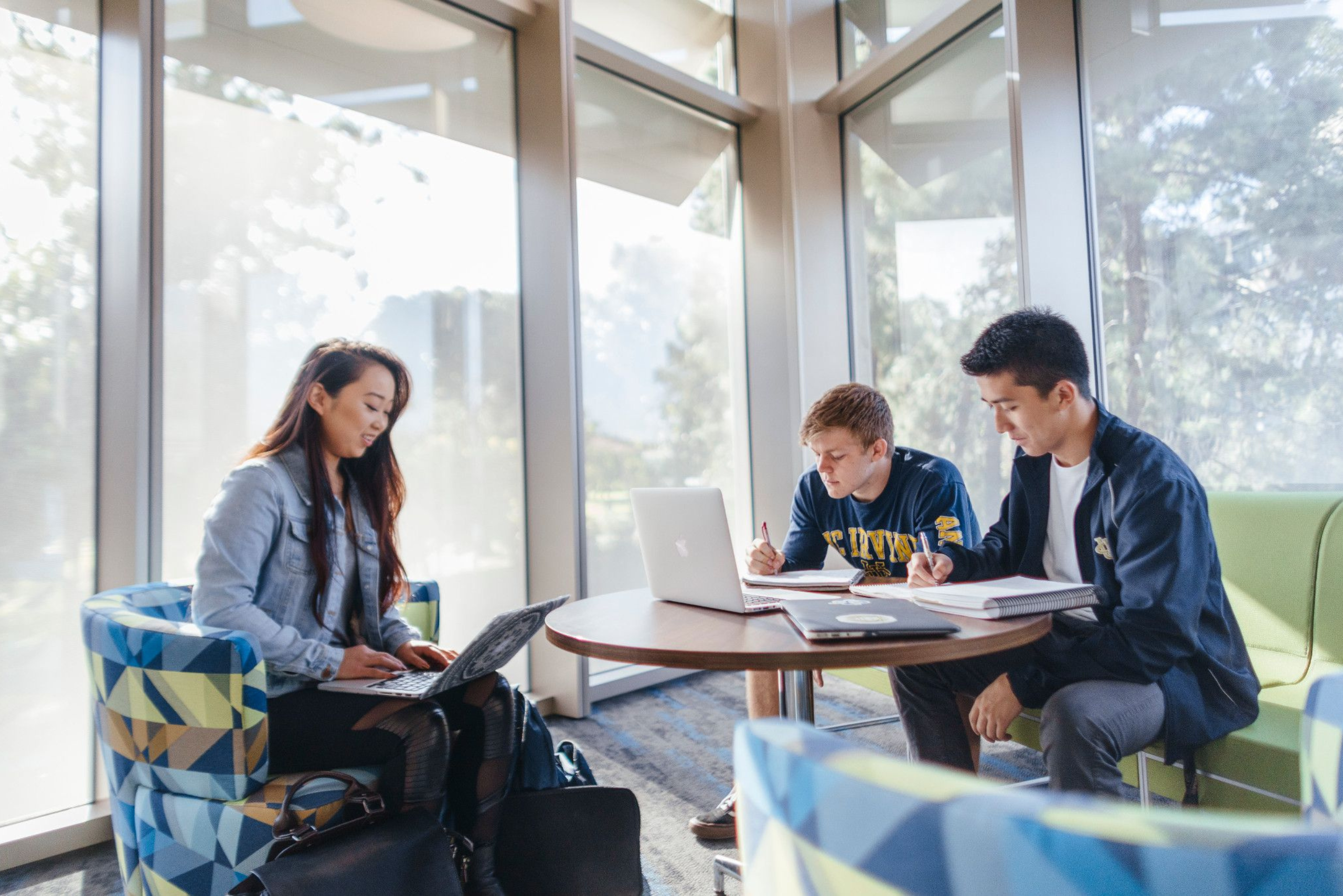 Three students study together in a bright campus lounge, using laptops and notebooks at a round table near large windows.