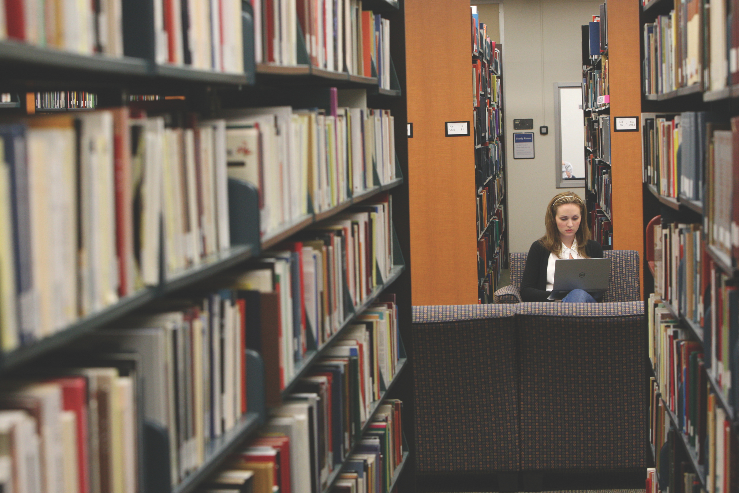 Person sitting with a laptop in a library, surrounded by tall bookshelves filled with books.