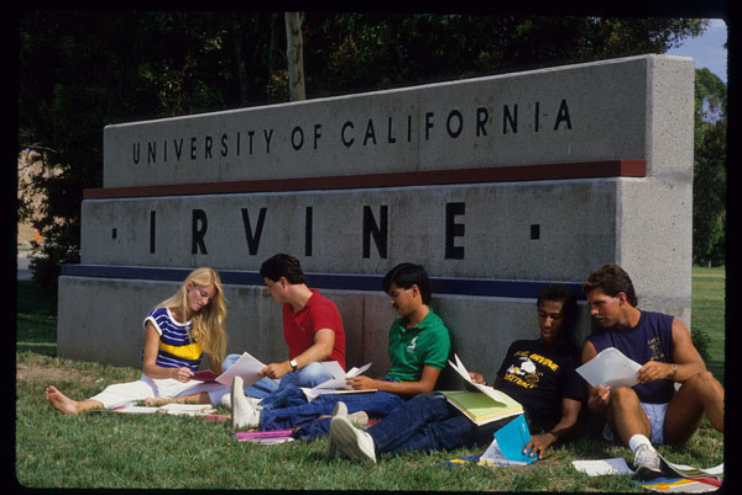 Students sit on the grass studying beside the University of California, Irvine campus entrance sign.