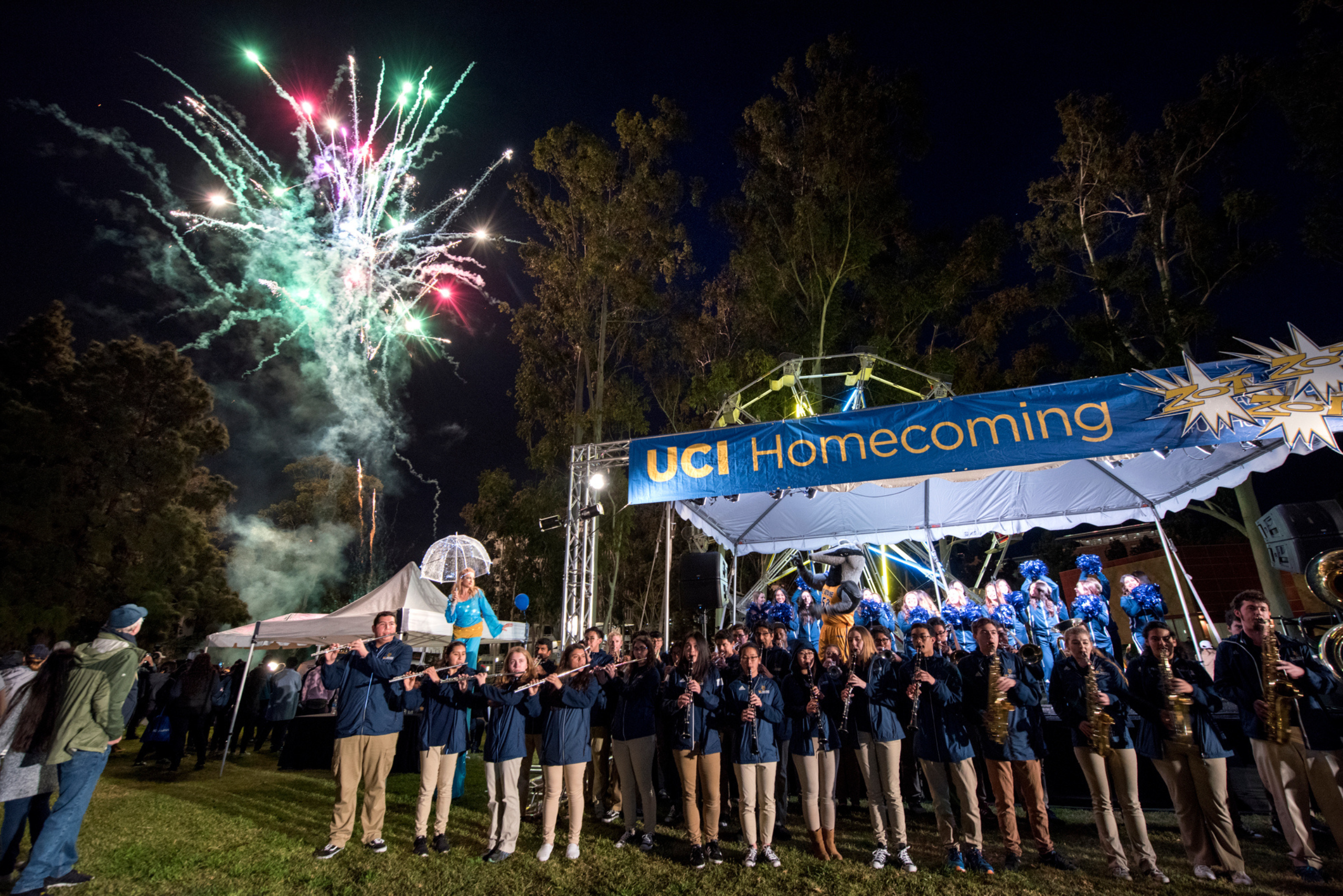 Fireworks light up the night sky above a UCI Homecoming celebration featuring a marching band and attendees gathered outdoors.