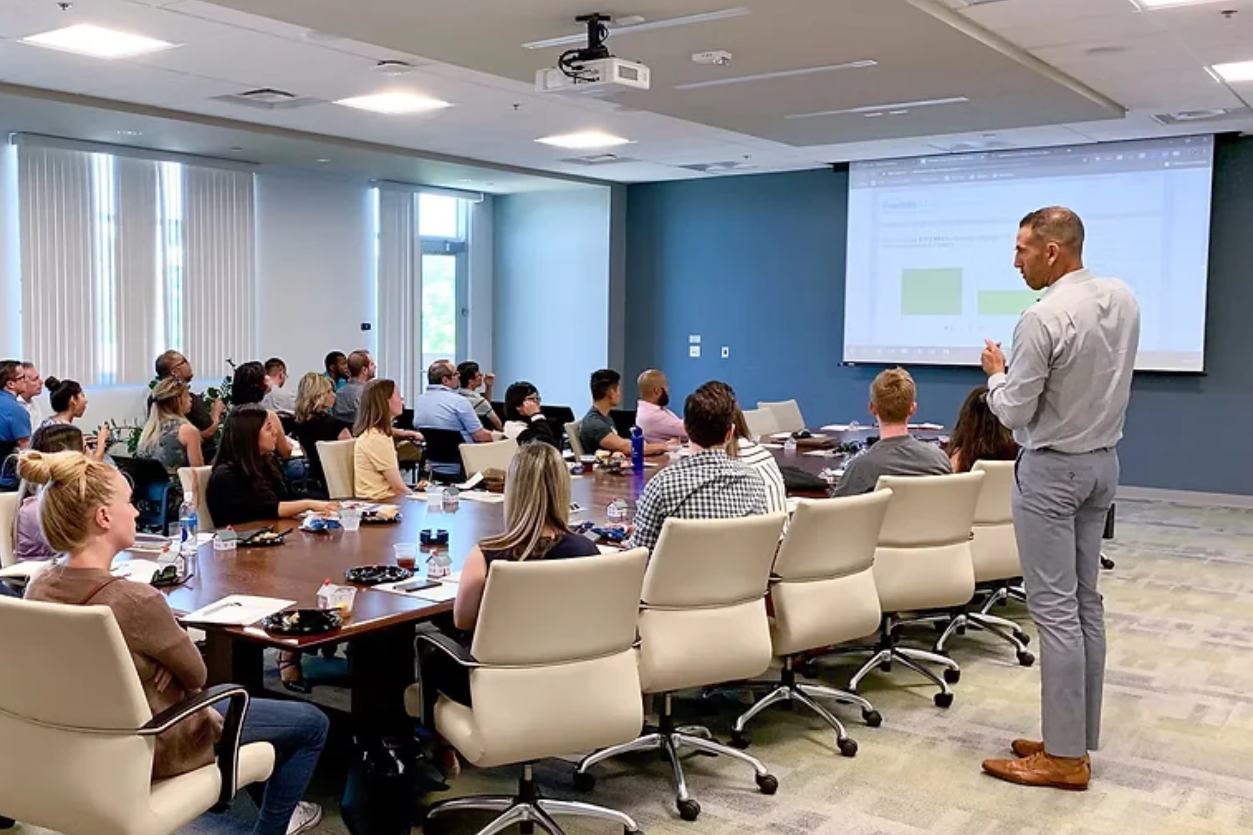 People seated around a large conference table attend a workshop in a modern meeting room while a presenter stands near a projected presentation screen at the front of the room.